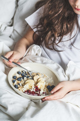 Beautiful little girl eating healthy oatmeal with fruits and berries in white bed.