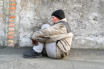 Dirty homeless woman sits under a gray wall