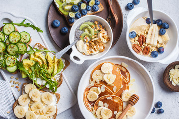 Flat lay of healthy vegetarian breakfast. Oatmeal with fruits, chia pudding, pancakes with banana and honey and toasts with fruits, vegetables and cream cheese.