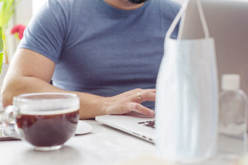 a man works at home with a laptop, a protective face mask, coffee and a sanitizer, quarantine due to the coronavirus pandemic