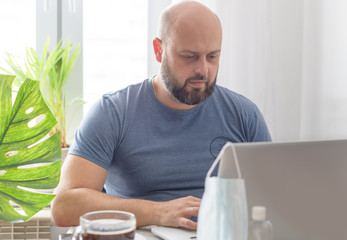 a man works at home with a laptop, a protective face mask, coffee and a sanitizer, quarantine due to the coronavirus pandemic