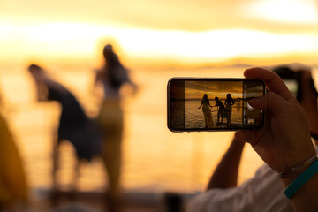 Close up of hand holding the phone to take picture of women enjoying beautiful sunset in front of yacht.