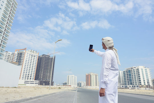 Middle east man wearing traditional white suit with head scarf taking picture of buildings with smartphone on city street over sky background, Bahrain.