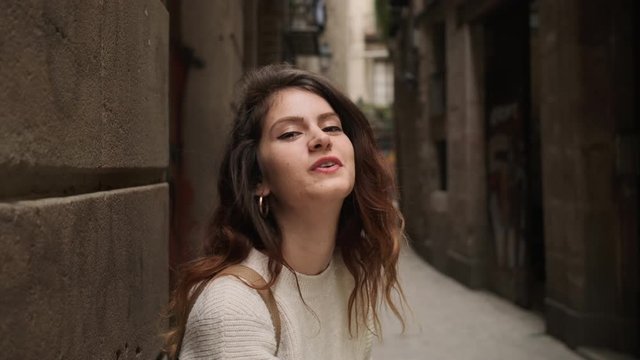 Tracking shot of charming hispanic girl happily looking in camera sending air kiss on narrow city street of Barcelona