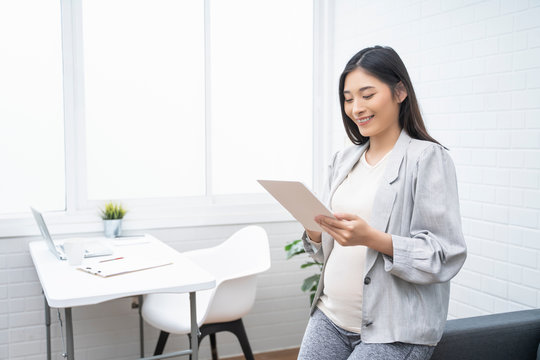 pregnant asian woman working from home in modern white office using smart tablet device with pen touching digital drawing notes, smiling with happiness of motherhood pregnancy expectant new born baby