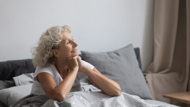 Peaceful calm positive middle aged senior retired woman sitting in bed after wakeup in weekend morning, enjoying good mood after good night rest relaxation, welcoming new day alone in bedroom.