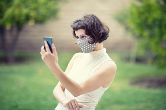 A White Woman In A Fabric Face Mask While Using Her Phone For Selfie And Video Chats. The New Recommendation - Everyone To Wear Masks In Public During The Coronavirus Covid-19 Pandemic.