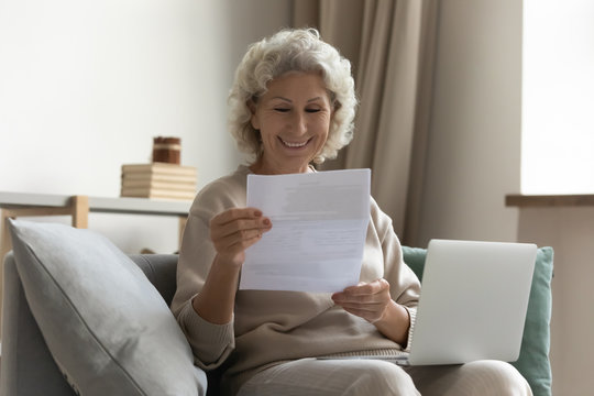 Happy Middle Aged Elderly Woman Sitting On Couch, Holding Computer On Knees Lap, Reading Paper Document Or Mail Letter From Friend. Smiling Mature Old Grandmother Excited By Good News Notification.