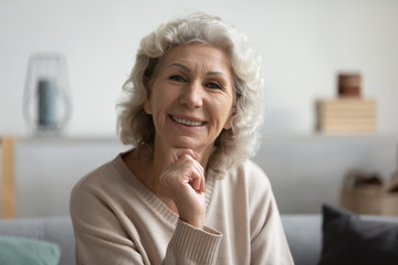 Head shot portrait of happy grey haired elderly mature woman touching chin. Smiling pleasant middle aged older grandmother looking at camera, enjoying free leisure weekend time alone at home.