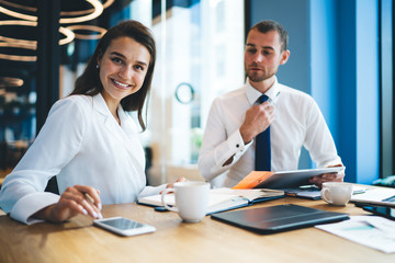 Portrait of cheerful female boss in formal clothing smiling at camera during cooperation brainstorming with handsome man with touch pad, happy successful colleagues teamwork on business project