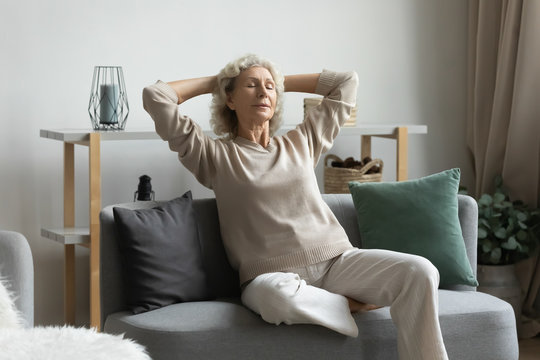 Calm peaceful middle aged mature hoary woman relaxing on cozy couch, enjoying lazy weekend time in living room, reducing stress, sleeping daydreaming napping breathing fresh air alone at home.