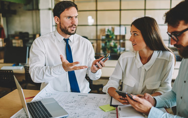 Fototapeta premium Caucasian employees feeling puzzled during cooperation brainstorming in office interior, young group of architects discussing problem of blueprint engineering collaborating at table with laptop
