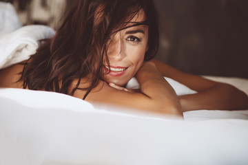 Obraz premium Close-up portrait Beautiful woman in the morning in a white bed after waking up, smiles and covers her face with hair. Soft selective focus. Toned photo.