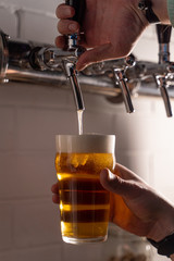 Man pouring light gold beer in a glass from a beer tap, pour with white beer foam, close up hands