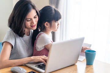 Asian young mother work from home. Woman use laptop for meeting with colleague while daughter sit on the lap and play. Mom happy to do job while taking care family. Covid-19, Social distancing concept