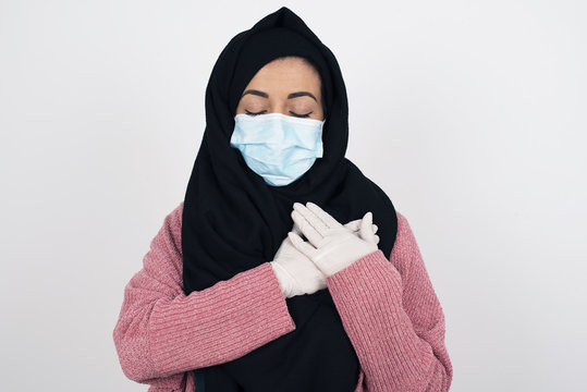 Faithful Muslim Woman Wearing Hijab And Face Mask Closes Eyes And Keeps Hands On Chest Near Heart, Shows Her Kindness Or Favour, Expresses Sincere Emotions, Being Kind Hearted And Honest.