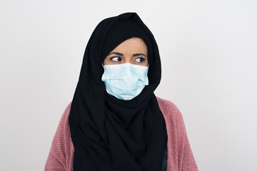 Naklejka premium Photo of amazed puzzled young muslim female wearing face mask adn hijab, curves lips and has worried look, sees something awful in front, isolated on white background.