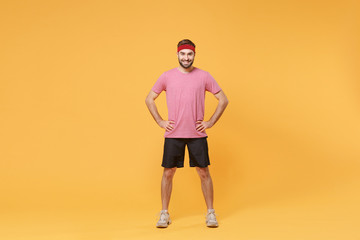 Fototapeta na wymiar Smiling young bearded fitness sporty guy sportsman in headband t-shirt spend weekend in home gym isolated on yellow background. Workout sport motivation concept. Standing with arms akimbo on waist.