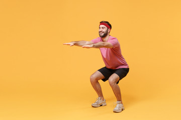 Smiling bearded fitness sporty guy sportsman in headband t-shirt spend weekend in home gym isolated on yellow background. Workout sport motivation concept. Doing squats exercising, spreading hands.