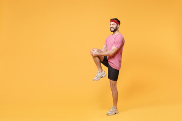 Fototapeta na wymiar Smiling young bearded fitness sporty guy 20s sportsman in headband t-shirt in home gym isolated on yellow background. Workout sport motivation concept. Doing warm up, stretching exercising for legs.