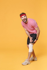 Ingured screaming young bearded fitness guy 20s sportsman in headband t-shirt in home gym isolated on yellow background. Workout sport motivation lifestyle concept. Touch knee with elastic bandage.
