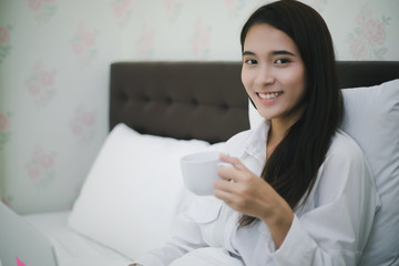 Asian woman is drinking a cup of coffee and using a notebook to work on the bed.She smiles and enjoys working at home.