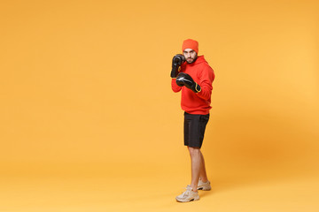 Fototapeta na wymiar Young bearded fitness sporty guy boxer sportsman in hat, hoodie, shorts in home gym isolated on yellow background. Workout sport motivation lifestyle concept. Doing boxing exercises in boxing gloves.