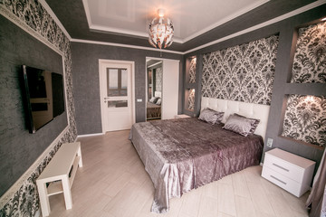 A grey bedroom with a bed and door. With chandelier, TV and bedside table