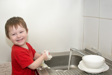 Happy little child in red clothes washes the dishes. Water flows from a faucet, the hands of a child in foam. Mom’s home chores