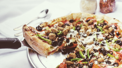 A view of a supreme style pizza pie, with a pie server lifting one of the cheesy slices, in a restaurant or kitchen setting.