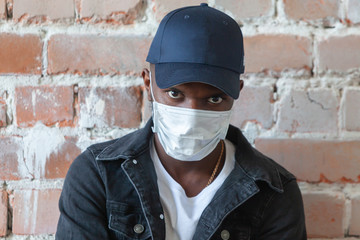 Young african black man wearing medical mask is standing in a cap with crossed arms shocked with coronavirus 2019 danger on background of ruined red brick wall