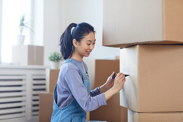 Side view portrait of smiling Asian woman writing on cardboard boxes labeling them for moving out...