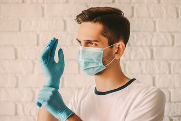 Young man hospital staff in medical mask on face wearing blue latex gloves on hands. Professional doctor surgeon in protective face mask putting on medical gloves. Coronavirus COVID-19 protection