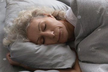 Close up head shot serene pleasant elderly senior woman lying under warm duvet on soft pillow, enjoying good night rest alone in bedroom. Peaceful middle aged grandma seeing sweet dreams in bed.