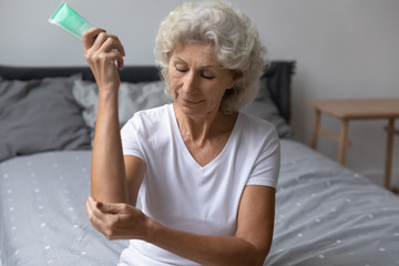 Pleasant happy middle aged senior hoary woman sitting on bed, applying moisturizing cream on elbow...