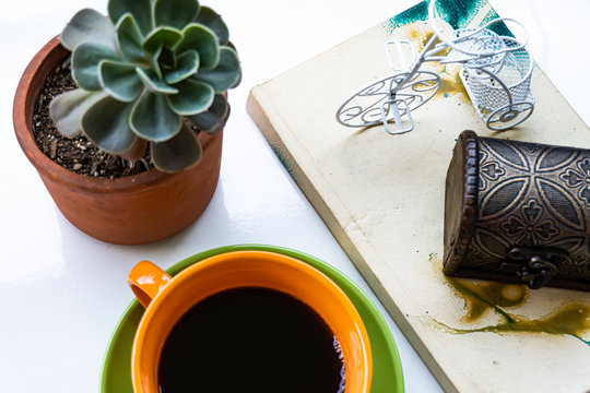 Taza de caf&eacute; con elementos decorativos, planta, libreta, bicicleta, ba&uacute;l de madera sobre una superficie blanca