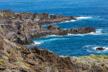 Fototapeta na wymiar Volcanic rock formation, cliffs of black lava on the rocky shore with crushing white waves over the Atlantic Ocean. Blue sky background. La Palma, Canary Islands.