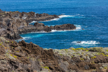 Volcanic rock formation, cliffs of black lava on the rocky shore with crushing white waves over the Atlantic Ocean. Blue sky background. La Palma, Canary Islands.
