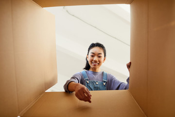 Low angle view at smiling Asian woman reaching into cardboard box while packing or unpacking for...