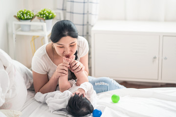 Asian young mother look at baby, hold son feet and kiss with warm love while cute boy close his eyes playing peekaboo with mom on bed at home. Mother child relationship is good for kid development.