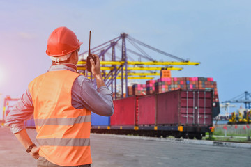 Engineer worker in port with walkie talkie in hand operation control Logistics and transportation of containers on train platform, with crane bridge, import, export industry and transport container