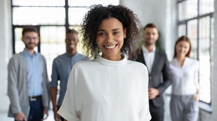 Close up headshot portrait of happy african american businesswoman looking at camera. Different smiling businesspeople standing behind of female company mentor. Leader of multi-ethnic team concept.