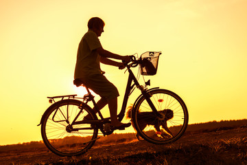 Senior woman riding bike and dog running in front, silhouette of riding person at sunset with pet
