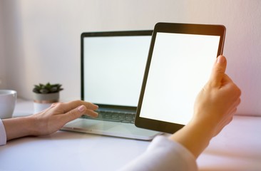 Close-up of tablet with blank screen in hands of young woman sitting at white table and touching screen. On table laptop with blank screen,cup of coffee and flowers. Minimalis. Mock up,space for ad