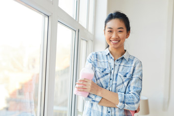 Waist up portrait of young Asian woman smiling at camera while washing windows and enjoying Spring cleaning, copy space