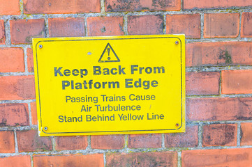 Sign at a railway platform warning passengers to keep back from the edge and to stand behind yellow line.