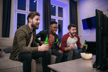 Likable joyful multiracial young guys cheering their favourite football team with screams and hands up during watching sport game on tv