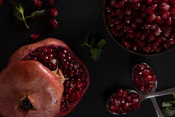 Ripe sliced pomegranate, a bowl and two spoons with pomegranate seeds on a black background. View...