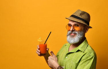 Grandpa in hat, green shirt, sunglasses. Twisting mustache, holding glass of fresh squeezed juice, posing on orange background