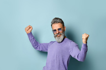 Gray-haired aged man in purple sweater, sunglasses. Dancing with clenched fist while posing on blue studio background. Close up